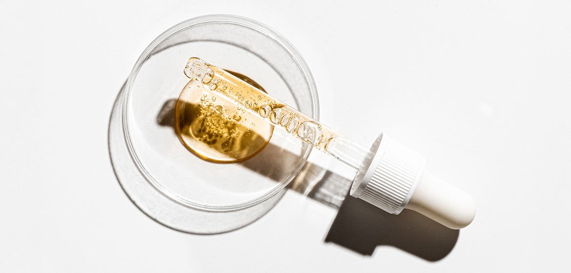 Eine Petrischale über der eine Pipette liegt vor einem weißen Hintergrund. In der Pipette und in Zentrum der Schale goldgelbe Flüssigkeit mit viele kleinen Bläschen.